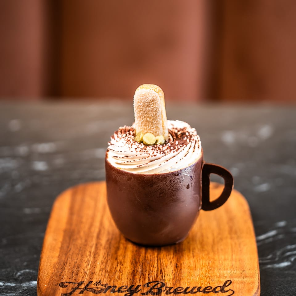 Chocolate mug topped with whipped cream, cocoa, and a toasted biscuit on a branded wooden serving board.