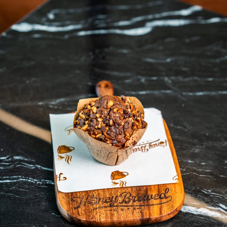 Chocolate muffin topped with chopped nuts and chocolate chips, served on branded paper atop a wooden café board.