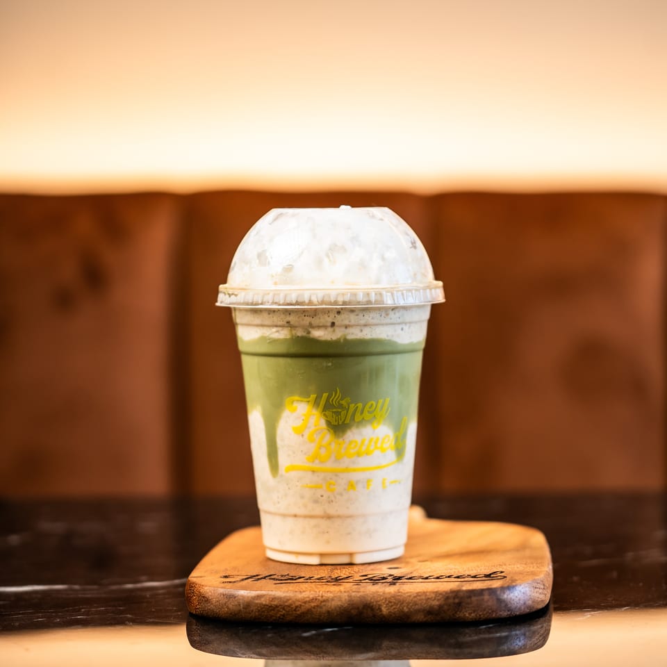 Layered green and cream iced drink with whipped topping in a Honey Brewed Café cup on a wooden board.
