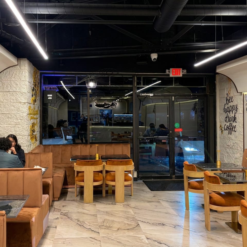 Modern café interior with tan booths, wooden chairs, marble tables, exposed ceiling ducts, and two seated patrons.