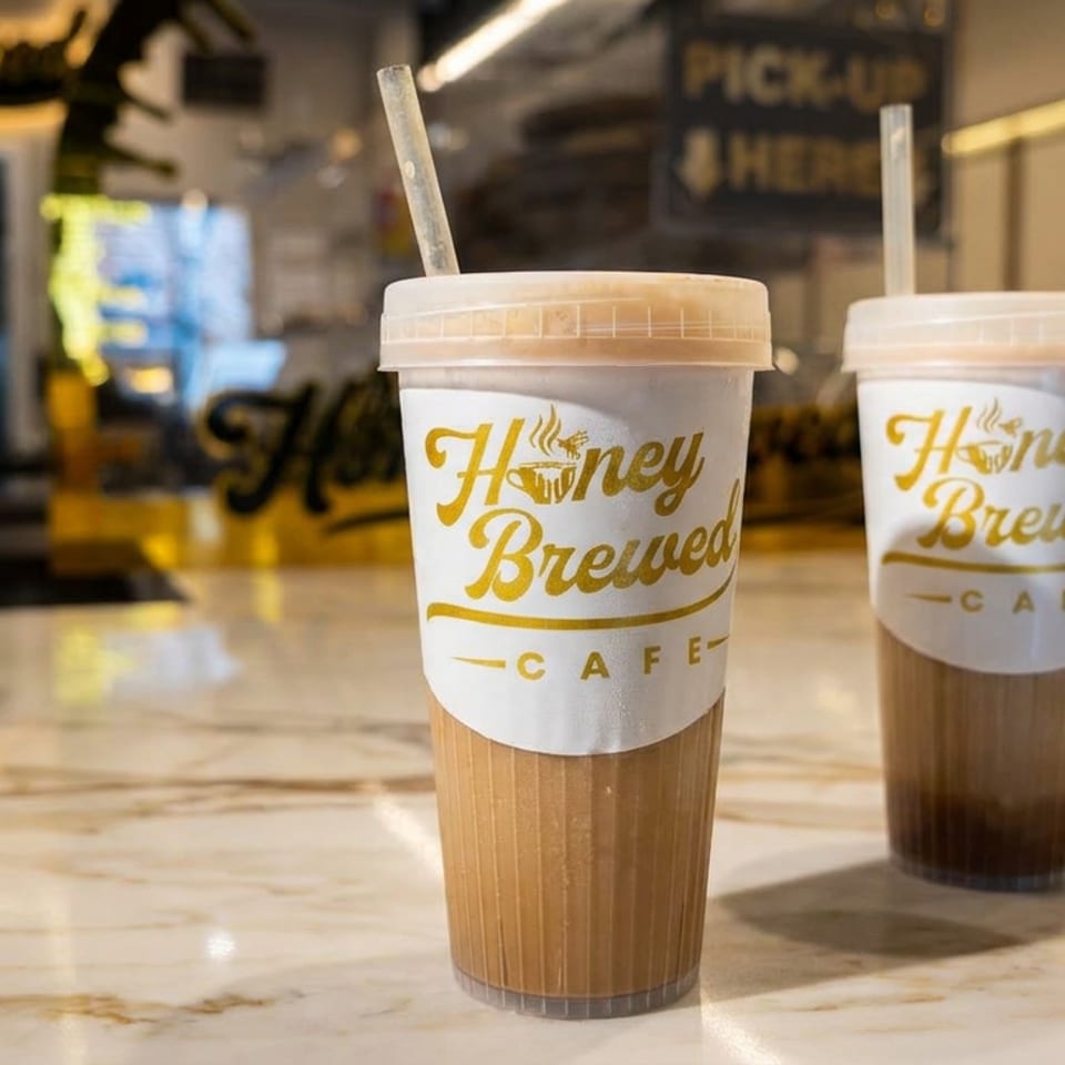 Two iced coffee drinks in Honey Brewed Cafe cups sit on a marble counter inside a warmly lit café.