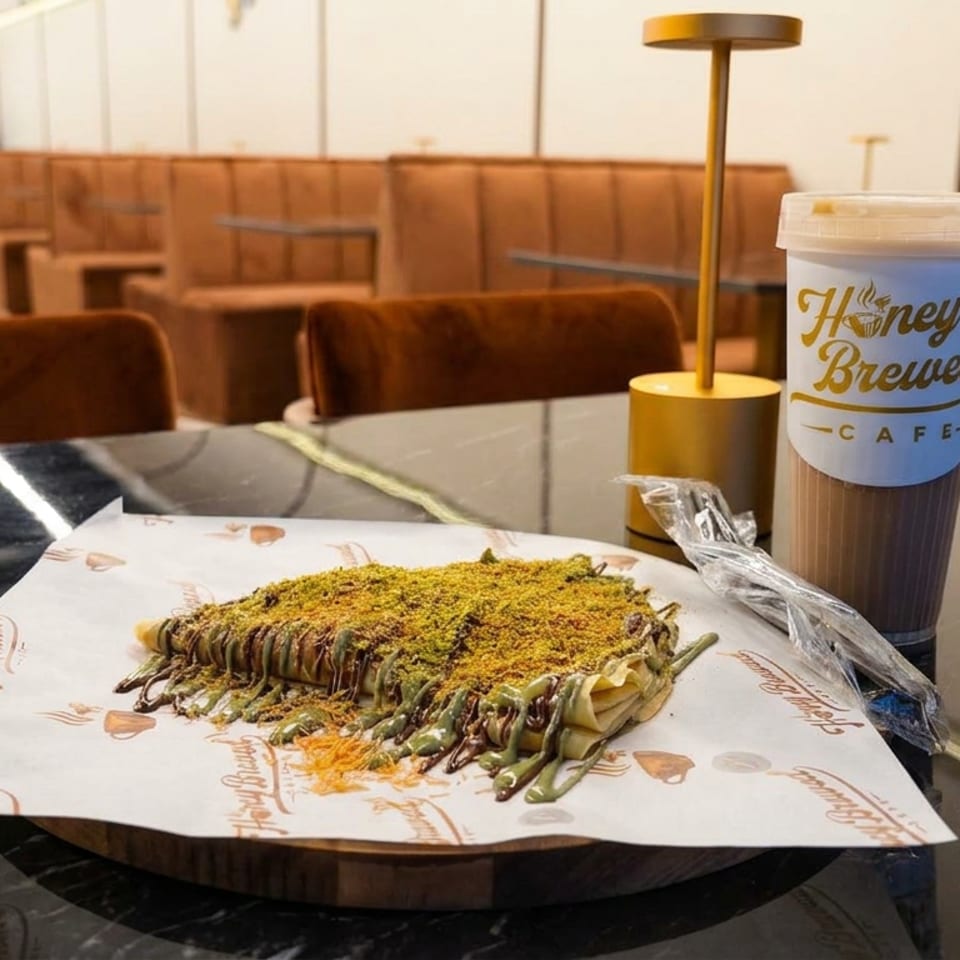 Pistachio-topped rectangular dessert drizzled with chocolate beside a Honey Brewed Cafe drink on a restaurant table.