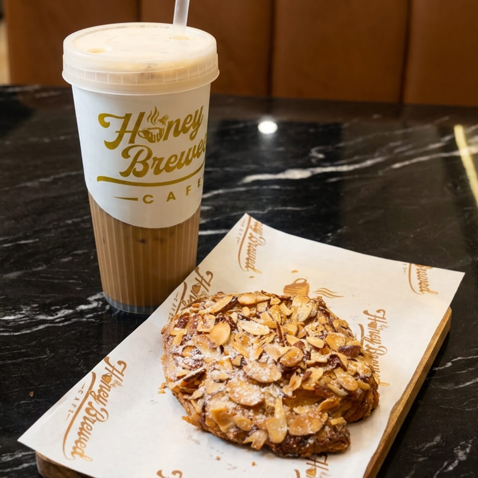 Iced coffee and an almond-topped pastry sit on branded paper atop a black marble table at Honey Brew Cafe.