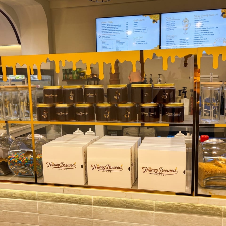 Honey Brewed Café counter displays jars, canisters, toppings, packaged goods, and illuminated digital menus behind a honey-themed shelf.