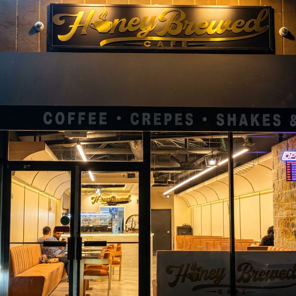 Honey Brewed Cafe storefront at night, with illuminated signage, open sign, and warmly lit seating visible through glass doors.