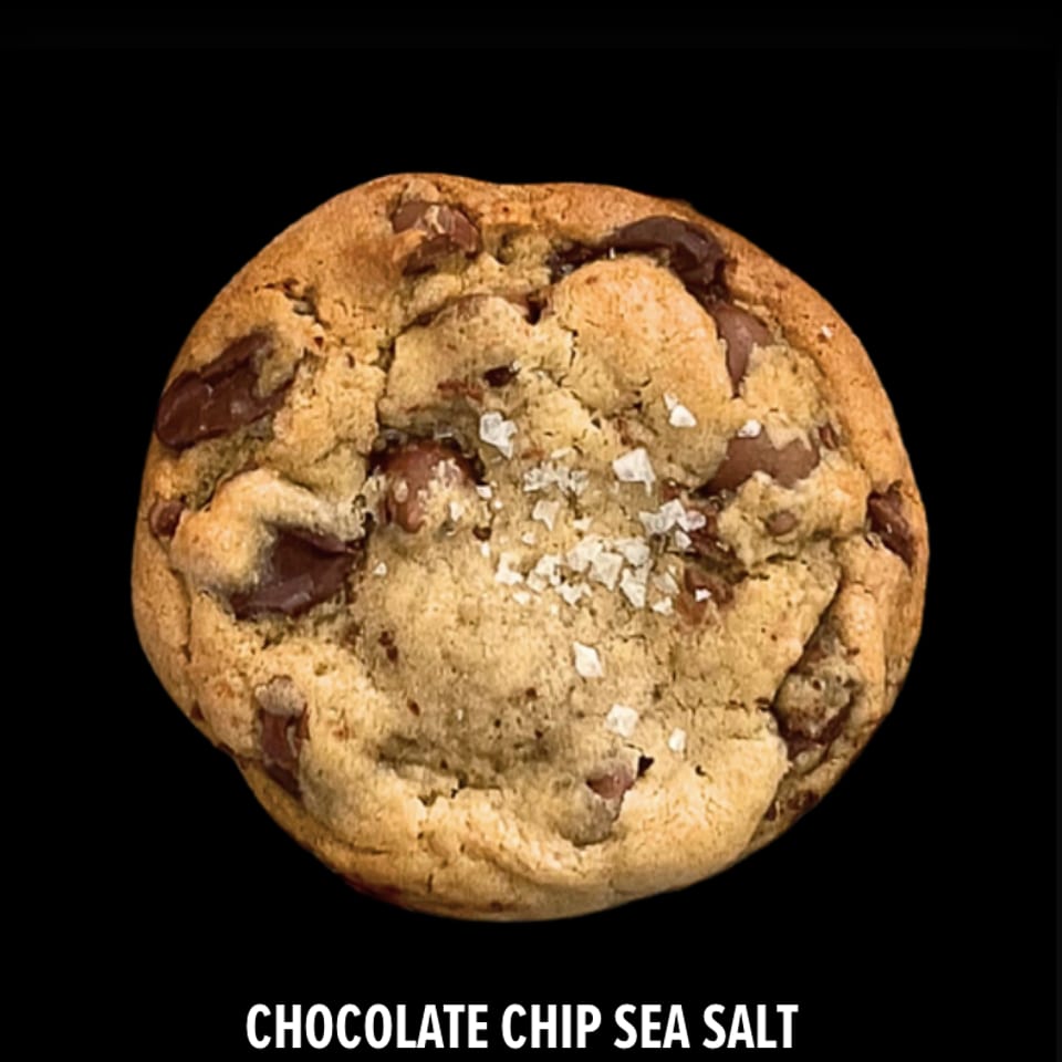 Large chocolate chip cookie topped with flaky sea salt against a black background.