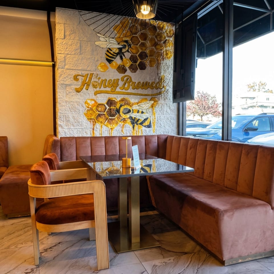 Cozy restaurant seating with brown leather booths, marble tables, large windows, and a honeybee mural reading “Honey Brewed.”
