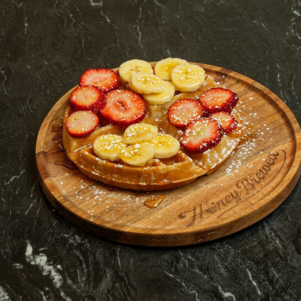 Belgian waffle topped with sliced strawberries and bananas, powdered sugar, and syrup on a wooden serving board.