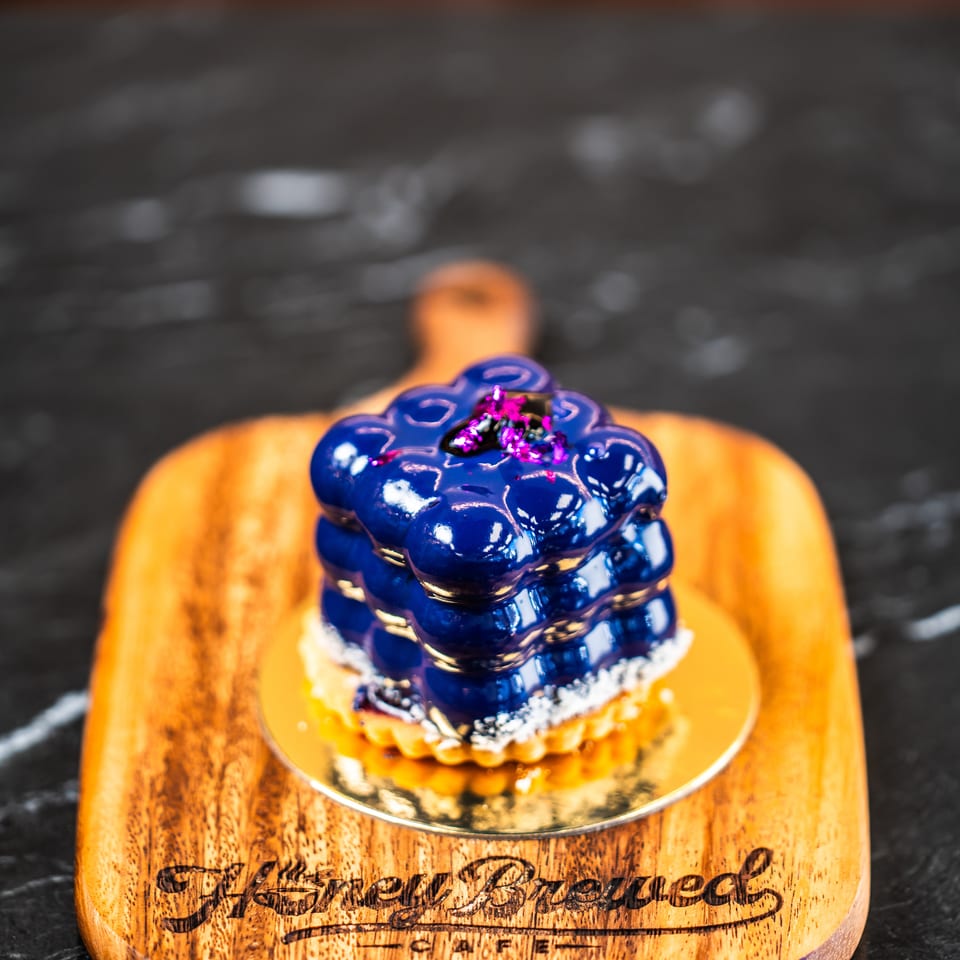 Blue glazed flower-shaped dessert topped with edible blossoms on a gold plate atop an engraved wooden serving board.