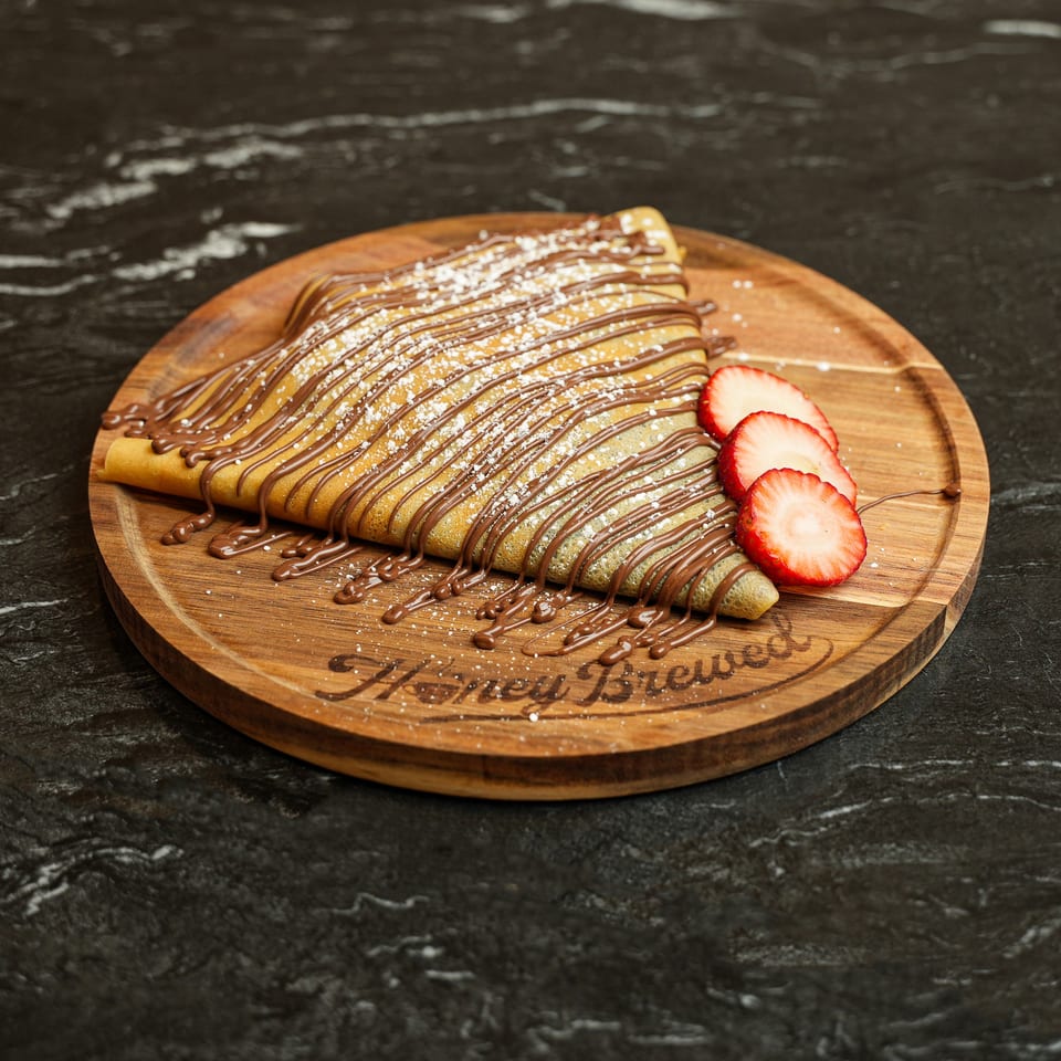 Golden crepe drizzled with chocolate and dusted with powdered sugar, served with sliced strawberries on a wooden platter.