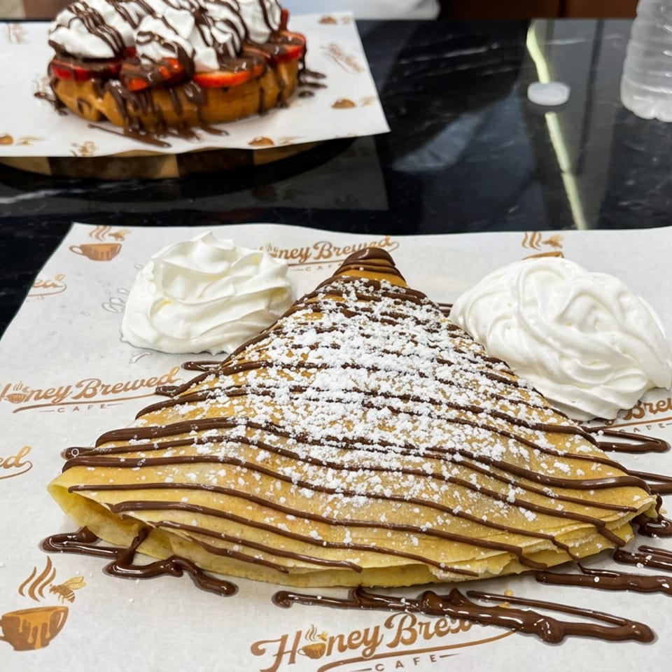 Triangular crepe drizzled with chocolate and powdered sugar, served with whipped cream at Honey Brewed Cafe.